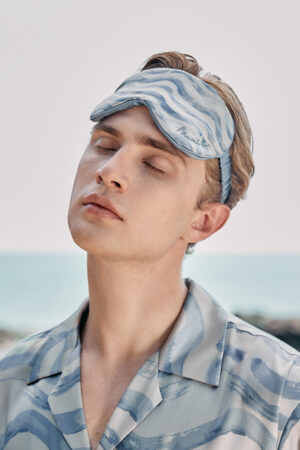 Close-up of the MANITO Ocean Wave Men's Silk Eye Mask worn by a man relaxing on rocks.