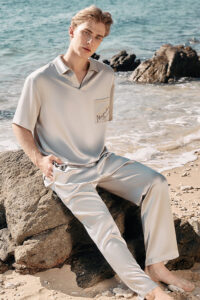 A man sits barefoot on a beach rock, wearing the MANITO Remington Men's Short Sleeve Pajama Set in ivory silk.