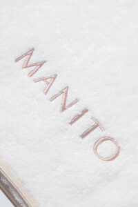 MANITO Soft Feeling 3 Pieces Bath Towel Set close-up of embroidered logo on white cotton towel with silk trim.
