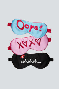 Three playful silk eye masks with embroidered designs; blue with 'Oops!', pink with 'XOXO', and black with 'shhhhhh...' on a neutral background.