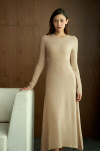 Woman wearing the Wunder Knit Dress in beige, standing by a sofa with a calm, elegant presence.