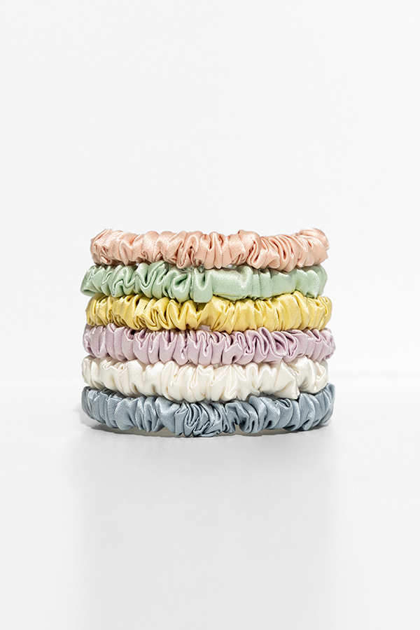 Stack of six skinny silk scrunchies in pastel shades of peach, yellow, green, purple, cream, and blue displayed on a bright white background.