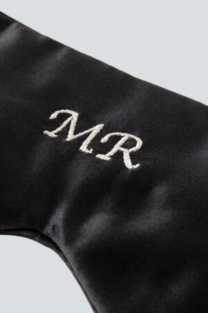 Close-up of a luxurious black silk eye mask with the monogram "MR" embroidered in elegant white script, highlighting the detailed craftsmanship.