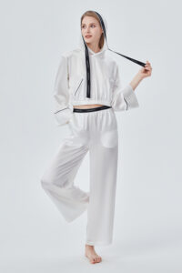 Woman wearing Sportif Silk Tracksuit in white, standing with one hand lifting logo drawstring, showcasing elastic waistband and relaxed fit.