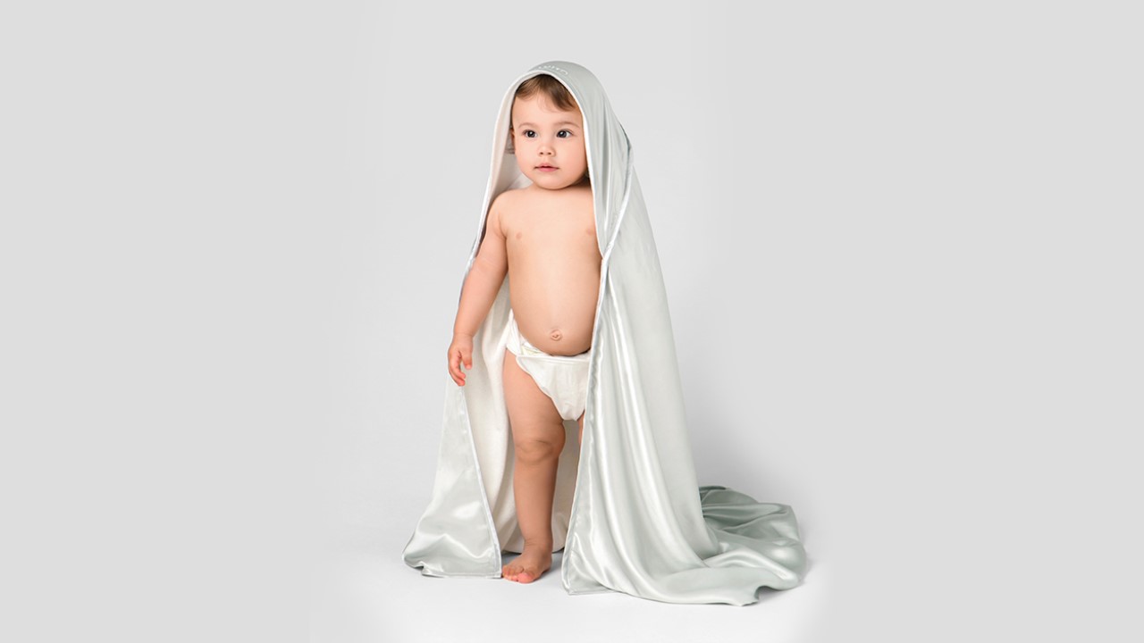 A baby stands centered in the image, covered with a silk sheet.