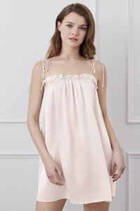 A woman wearing a pastel pink Blooming Short Slip Dress with delicate shoulder ties and scalloped neckline detail.