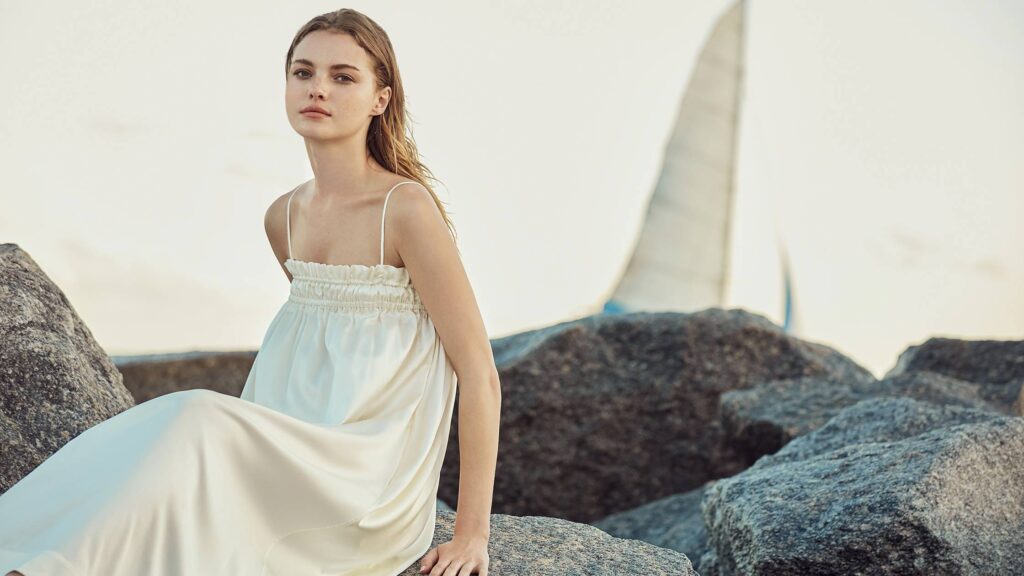Woman perched on rocks by the sea wearing a MANITO Silk Nightgown, epitomizing elegance and comfort.