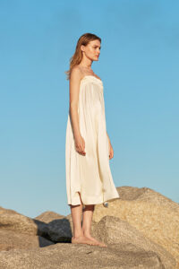 Elegantly poised woman in Blooming Silk Midi Slip Dress showcasing a shirred bodice and flowy skirt on a rocky shore.