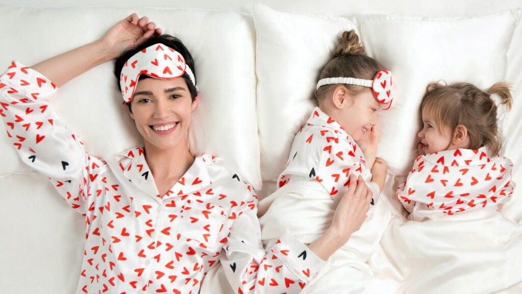 A woman and two young children in matching white pajamas with red heart patterns, lying in bed with a white sleeping mask on their foreheads, smiling and enjoying a cozy family moment.