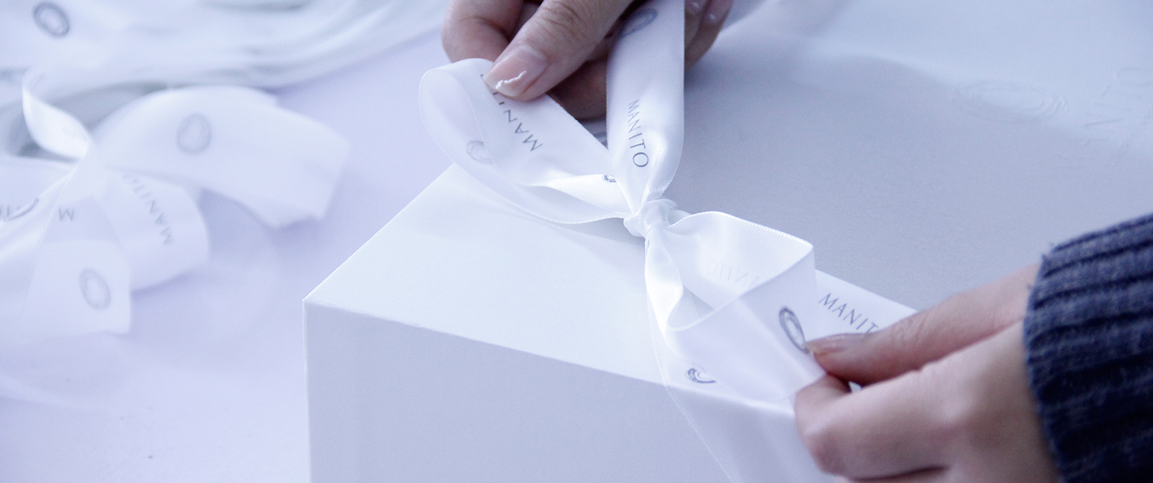 Hands tying a satin ribbon on MANITO's pristine white gift box, symbolizing attention to detail in presentation