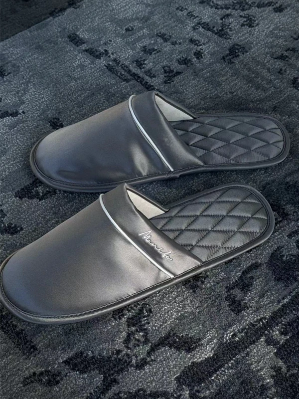 Pair of MANITO black silk slippers with quilted lining and embroidered logo, placed on a dark textured rug.
