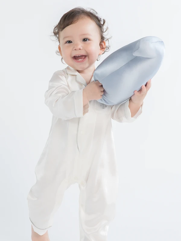 Baby wearing MANITO white silk romper, holding a blue silk pillow with a joyful smile.