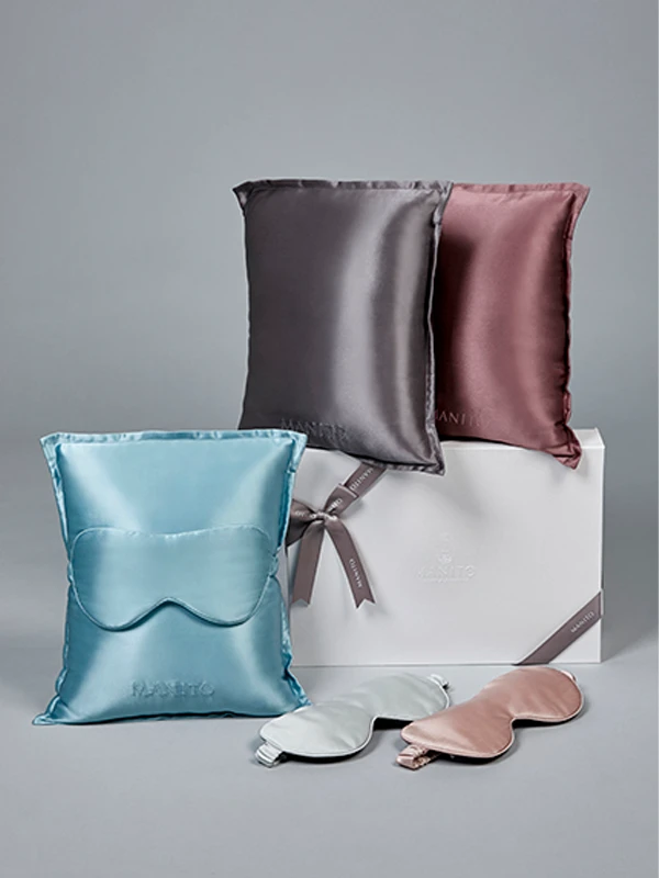 MANITO silk pillowcase and eye mask gift set in assorted colors, styled with gift box and ribbon on a grey background.