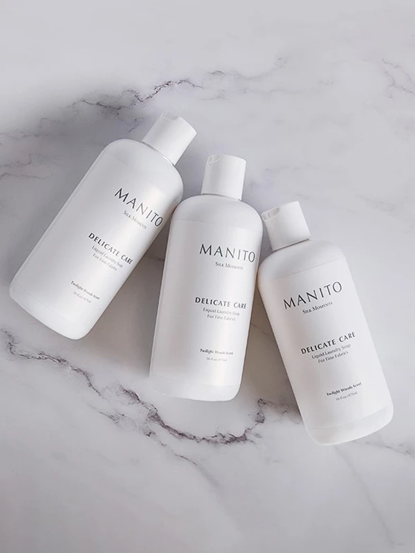 Three bottles of MANITO Delicate Care liquid laundry soap for silk fabrics, styled on a marble surface.