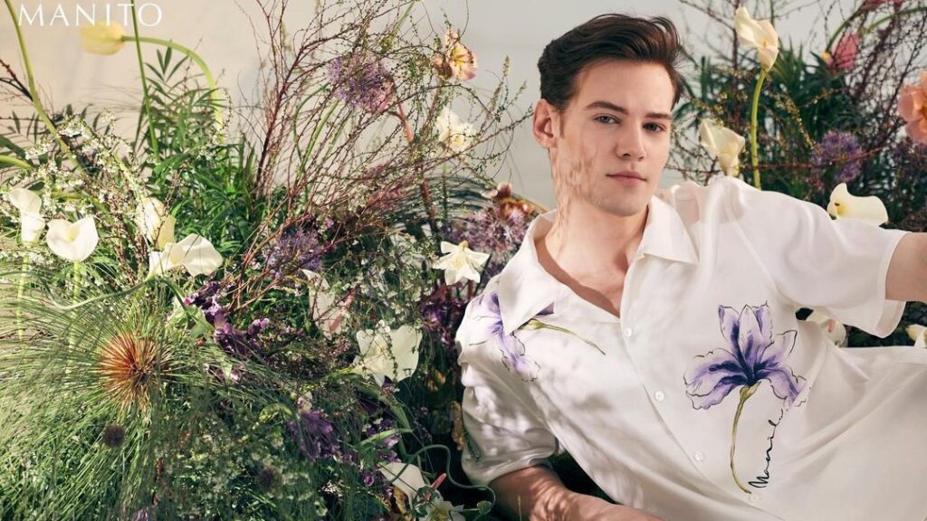 A young man in a men's short sleeve pajama set with a large purple iris print, reclining elegantly amid a lush display of wildflowers.