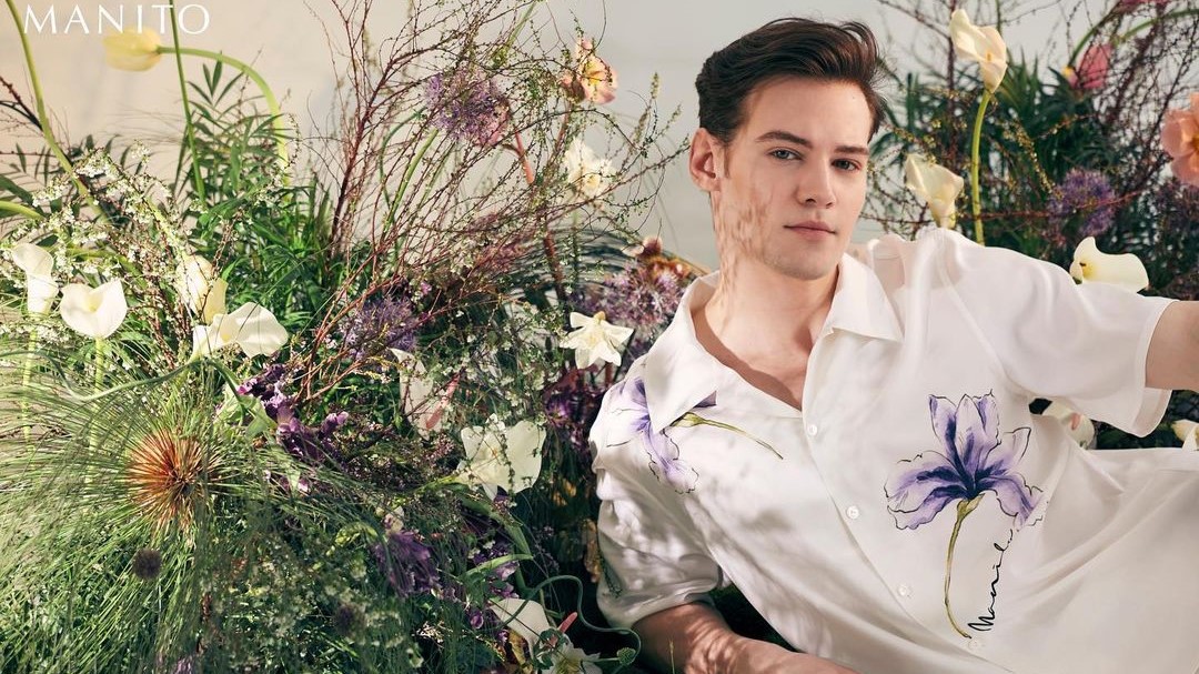 A young man in a men's short sleeve pajama set with a large purple iris print, reclining elegantly amid a lush display of wildflowers.