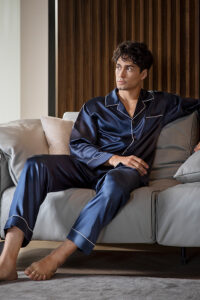 Man lounging in an Authentic Navy Silk Pajama Set with white piping detail, sitting on a sofa.