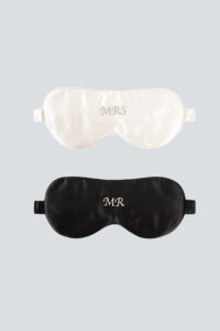 Two silk eye masks with monograms, one in ivory white with "MRS" and the other in black with "MR", set against a light gray background.