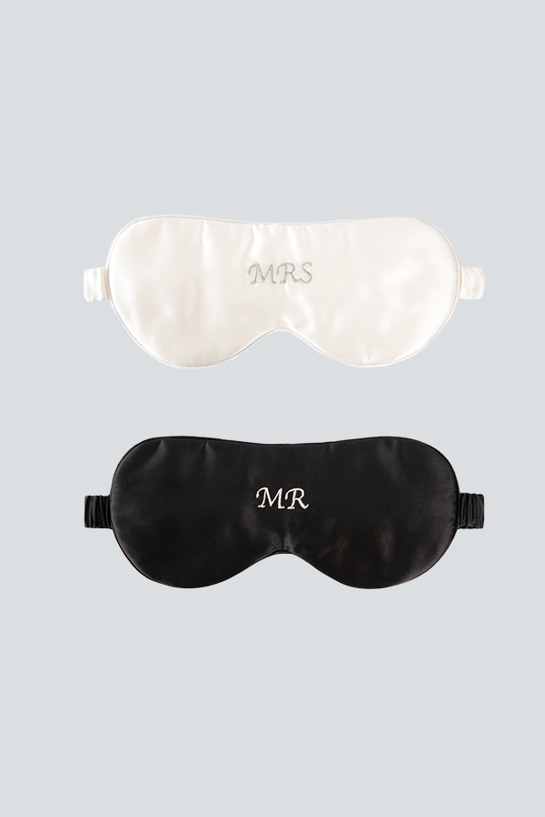 Two silk eye masks with monograms, one in ivory white with "MRS" and the other in black with "MR", set against a light gray background.