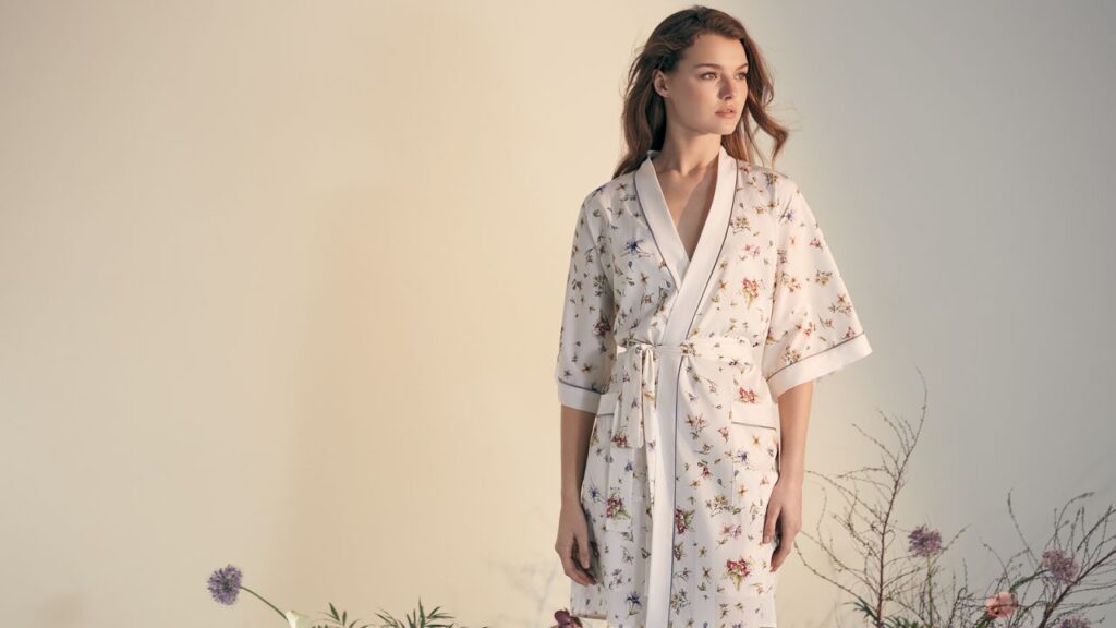 A woman in a light floral mulberry silk robe poses with soft elegance amidst a serene botanical setting.