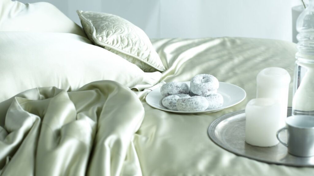 Close-up of silk bed sheets with powdered donuts on a plate and candles on a tray.