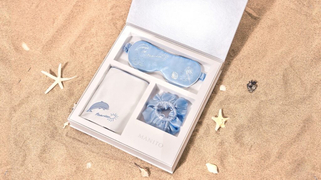 A blue silk travel set with an embroidered eye mask, scrunchie, and pillowcase displayed on a sandy beach.