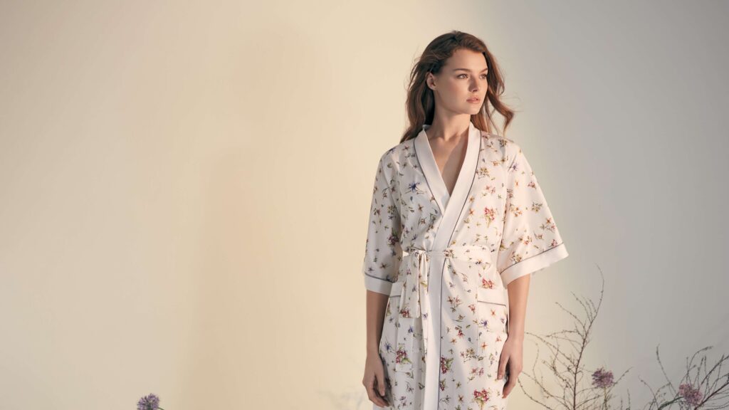 Woman wearing a floral patterned white silk kimono pajama standing against a light background.