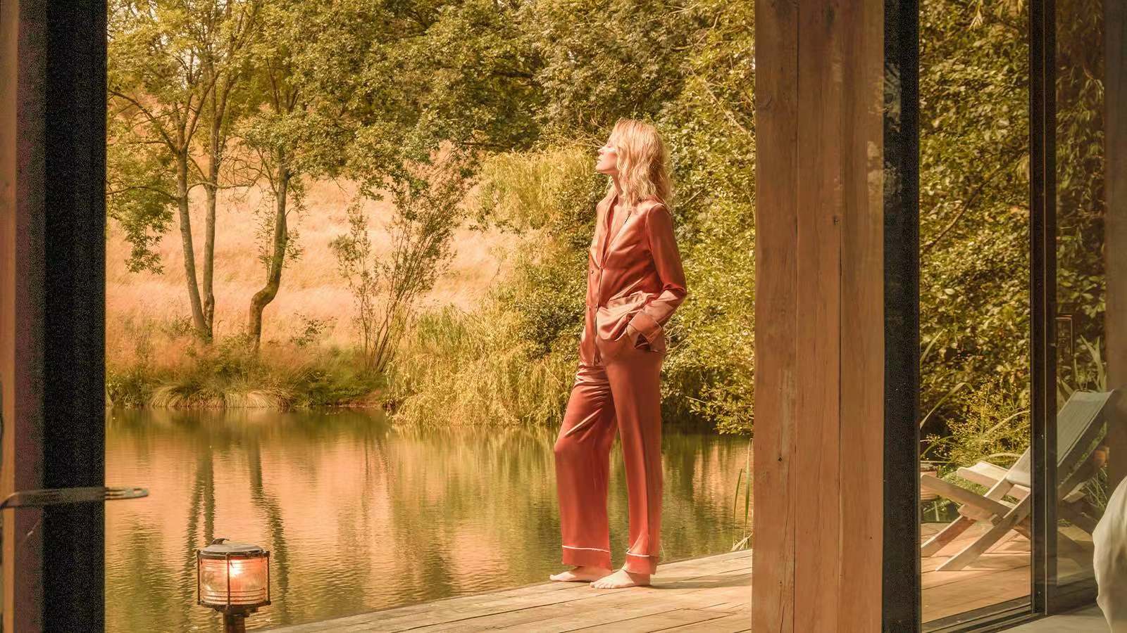Reflecting on the care of luxury fabrics, a woman in a mulberry silk loungewear gazes thoughtfully over a tranquil river, considering the best methods to maintain silk's sheen.