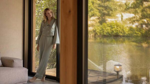 A woman in a mulberry silk pajama set stands by the curtain, peering out to a serene lakeside view, contemplating if you can steam silk to remove wrinkles.