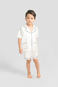 Happy young boy in white silk short pajama set with black piping detail.