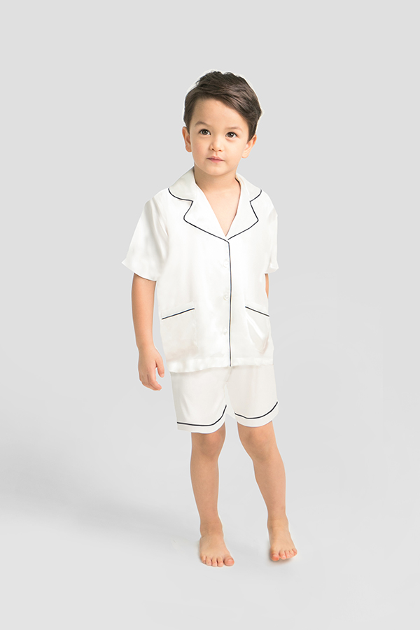 Happy young boy in white silk short pajama set with black piping detail.