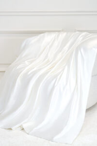 Gleam Silk Quilt in soft white silk draped over a chair, catching gentle light for a cozy bedroom mood.