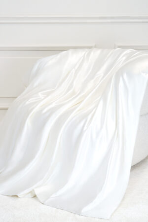 Gleam Silk Quilt in soft white silk draped over a chair, catching gentle light for a cozy bedroom mood.