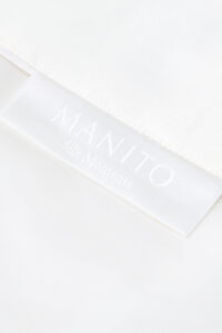 Corner of Gleam Silk Quilt showing the MANITO Silk Moments label and fine stitching on crisp white silk.